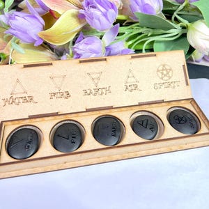 May include: A wooden box with five black stones, each engraved with a symbol and a word: Water, Fire, Earth, Air, and Spirit. The box is open, revealing the stones. The background includes purple flowers.