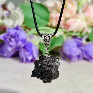Pode incluir: Um pingente mineral castanho-escuro com textura rugosa suspenso por um cordão preto. O pingente apresenta uma estrutura cristalina complexa e multifacetada presa a um passador de metal prateado com um intrincado padrão de volutas em relevo.