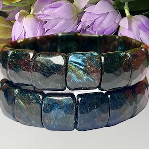May include: Two stacked bracelets made of polished, square-shaped dark blue and black gemstone beads. The stones feature iridescent, chatoyant streaks of metallic blue and brown, creating a reflective, multi-dimensional surface texture.