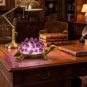 Può includere: Una statuetta decorativa a forma di tartaruga con un guscio composto da molteplici punte di cristallo di ametista viola. La tartaruga ha un corpo e una testa in metallo color oro, con il gruppo di cristalli illuminato dall'interno.