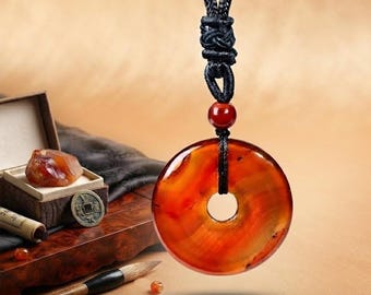 May include: A circular, translucent orange and red carnelian pendant suspended from a black braided cord. The pendant features a central hole and is accented by a small, round, dark red bead positioned above the main stone on the cord.