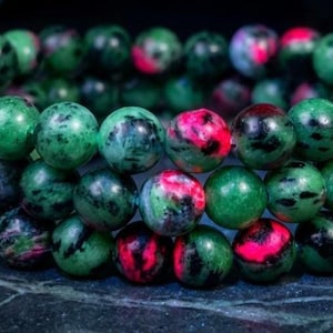 May include: A beaded bracelet composed of polished, spherical stones featuring a mottled pattern of deep forest green, vibrant magenta pink, and dark black inclusions. The beads are strung together in a circular arrangement, creating a continuous loop.