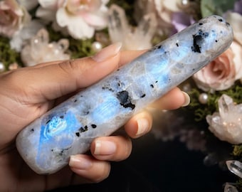 Puede incluir: Una varita cilíndrica de piedra lunar pulida con una superficie lisa y semitranslúcida, destellos azules iridiscentes e inclusiones minerales oscuras dispersas por toda la piedra, mostrando una textura cristalina natural y brillo.