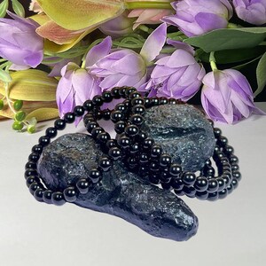 May include: A strand of polished black spherical beads arranged in a circular loop, resting upon a dark, textured, irregular stone surface. The beads have a glossy, reflective finish, and the stone displays a deep, dark, and pitted appearance.