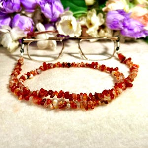 May include: A pair of eyeglasses with a gold-toned metal frame attached to a beaded chain made of small, irregular, polished carnelian stones in shades of orange, red, and translucent white, creating a vibrant and textured accessory for eyewear.