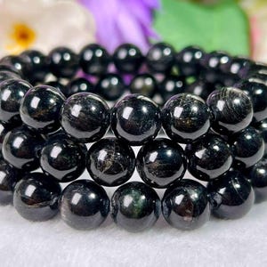 Puede incluir: Una pulsera de tres hilos compuesta por cuentas esféricas negras pulidas. Cada cuenta presenta una superficie lisa y reflectante con sutiles inclusiones fibrosas oscuras visibles dentro de la piedra, ensartadas en un cordón elástico flexible.