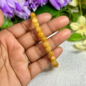 May include: A beaded bracelet featuring golden-yellow, round beads. The bracelet is held in a hand, with a silver ring visible on a finger. The background includes purple and yellow flowers.