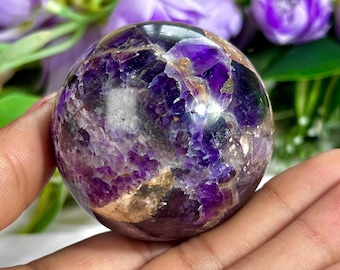Puede incluir: Una esfera de amatista pulida, que muestra una mezcla de tonos púrpura intenso y blanco translúcido. La superficie de la gema presenta patrones intrincados e inclusiones naturales, creando una textura visual única. Esta esfera decorativa es una hermosa adición a cualquier colección de cristales.