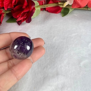 May include: A polished, oval-shaped amethyst stone, showcasing a gradient of deep purple to translucent hues. The gemstone is held in a hand, with a backdrop of red roses and greenery.