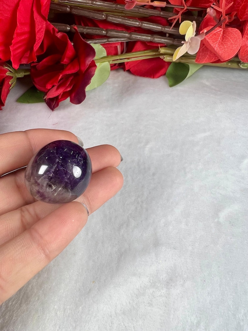 May include: A polished, oval-shaped amethyst stone, showcasing a gradient of deep purple to translucent hues. The gemstone is held in a hand, with a backdrop of red roses and greenery.
