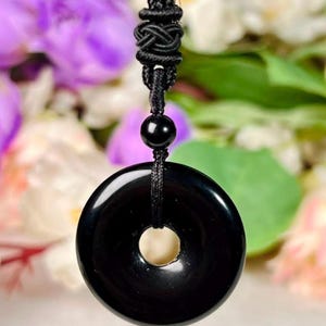 Puede incluir: Un colgante circular negro con un acabado liso y pulido y un orificio central, suspendido de un cordón negro. El cordón presenta un nudo decorativo sobre una cuenta negra individual, proporcionando una sujeción segura para la pieza de piedra.