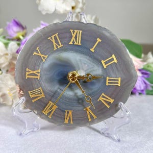 May include: A circular gray agate slice clock featuring gold-colored Roman numerals and matching gold-colored clock hands. The natural stone face displays concentric banding patterns in shades of blue and gray, mounted on a clear plastic stand.