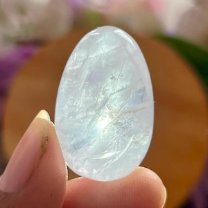May include: A polished, egg-shaped gemstone with a translucent, pale blue appearance. The stone features internal fractures and inclusions that catch the light, creating a shimmering effect across its smooth, rounded surface.