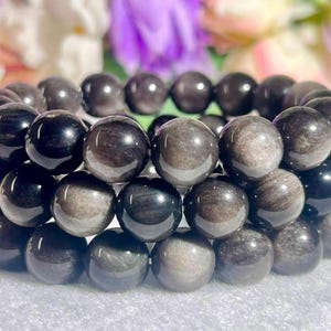 May include: A triple-strand bracelet composed of polished, spherical obsidian beads. The beads exhibit a smooth, reflective surface with a mix of deep black and shimmering silver-grey metallic tones, arranged in a uniform, circular elastic configuration.