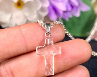 May include: A clear, translucent cross pendant made of polished stone or glass, suspended from a silver-toned metal chain. The cross features a smooth, rounded surface with visible internal textures and is attached to a simple metal bail.