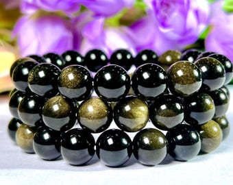 Könnte beinhalten: Ein Armband aus polierten, runden, schwarzen Perlen mit goldenem Schimmer. Die Perlen sind in drei Reihen gestapelt, wodurch ein geschichteter Effekt entsteht. Im Hintergrund sind verschwommene lila Blumen zu sehen.