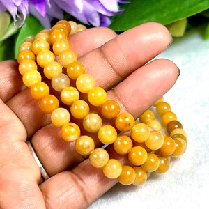 May include: Three strands of yellow-orange beaded bracelets. The beads are round and have a polished finish. The bracelets are displayed against a soft background with hints of purple and green.