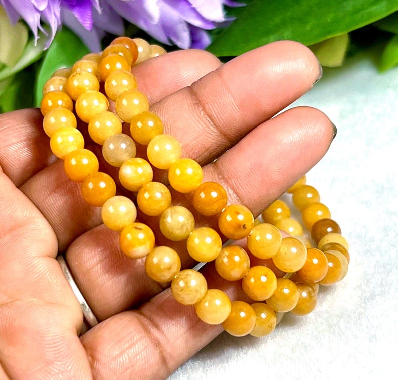May include: Three strands of yellow-orange beaded bracelets. The beads are round and have a polished finish. The bracelets are displayed against a soft background with hints of purple and green.