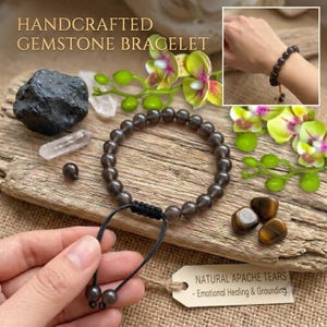 May include: A bracelet made of round, translucent dark gray gemstone beads with a black adjustable cord closure. The bracelet is accompanied by a tag labeled NATURAL APACHE TEARS - Emotional Healing & Grounding.