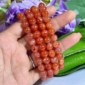 May include: A strand of round, reddish-orange gemstone beads. The beads are held in a hand, with purple flowers and green leaves in the background. The beads have a polished, glossy finish.