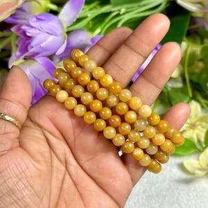 May include: Several strands of yellow-orange gemstone beads. The beads are round and appear to be strung together to form bracelets. The background includes purple flowers and green foliage.