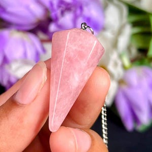 May include: A faceted, cone-shaped pink quartz pendant with a smooth, polished surface. The stone features a light pink hue with subtle white marbling throughout. A small silver-toned metal bail is attached to the top, connecting to a silver chain.