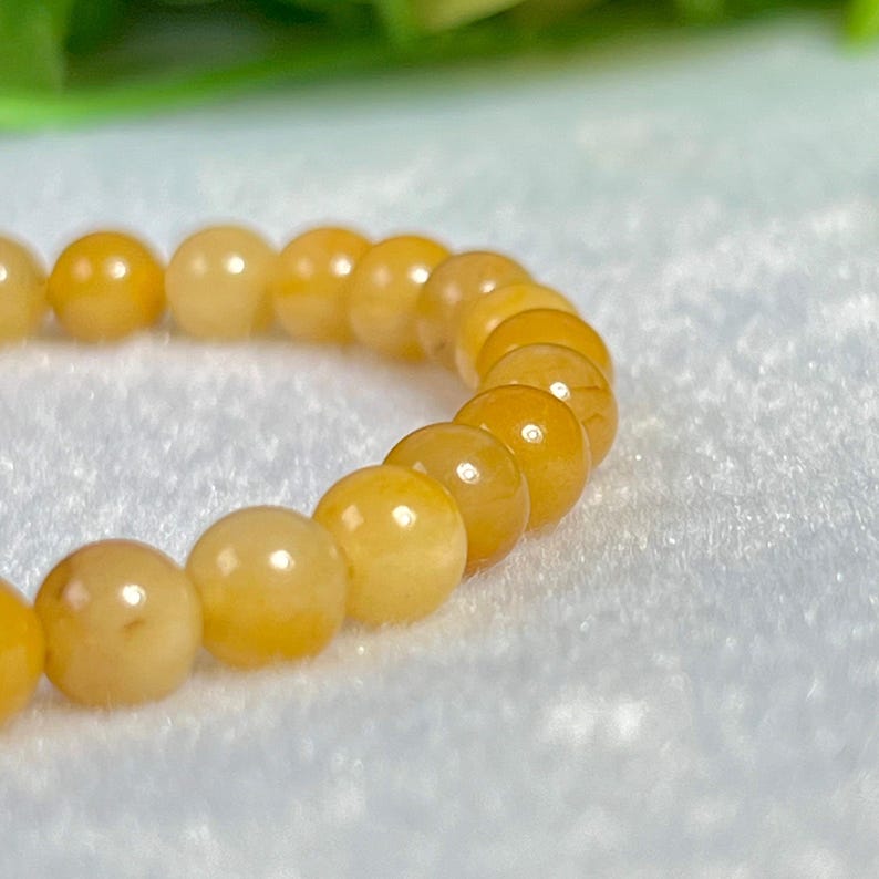 May include: A beaded bracelet featuring round, amber-colored beads. The beads have a smooth, polished surface and are strung together to form a complete circle. The bracelet is displayed on a white, textured surface.