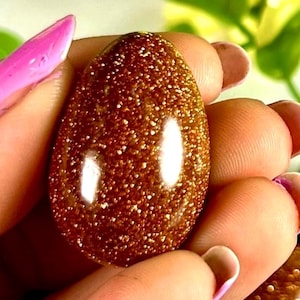 Puede incluir: Una gema pulida de forma ovalada con un aspecto marrón dorado brillante. La piedra tiene una textura moteada y un acabado brillante, que refleja la luz. La gema se sostiene en una mano con esmalte de uñas rosa.
