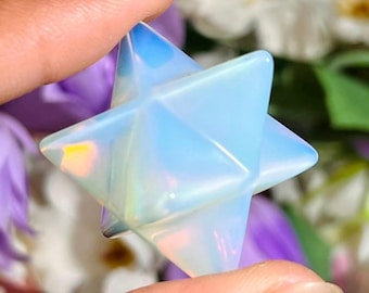 Puede incluir: Un cristal translúcido en forma de estrella, de color blanco lechoso con un brillo iridiscente. El objeto presenta múltiples facetas triangulares que forman una figura geométrica Merkaba, mostrando reflejos de luz azul y naranja.