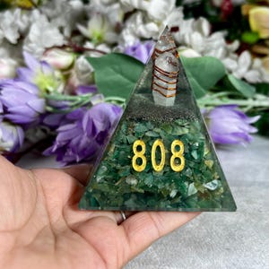 May include: A green pyramid-shaped resin art piece with the number "808" in gold. The pyramid contains green stones and a clear quartz crystal wrapped in copper wire. The background features purple flowers.