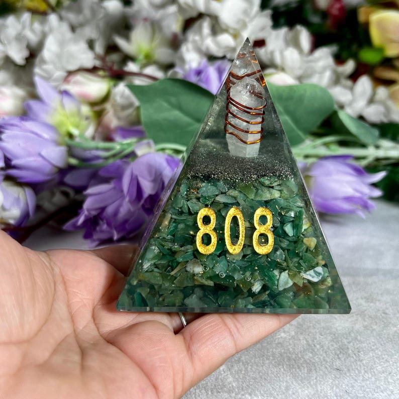 May include: A green pyramid-shaped resin art piece with the number "808" in gold. The pyramid contains green stones and a clear quartz crystal wrapped in copper wire. The background features purple flowers.