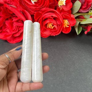 Puede incluir: Dos varitas de cristal cil&iacute;ndricas blancas con aspecto veteado. Las varitas se sostienen en una mano, con un fondo de vibrantes rosas rojas. Las varitas son lisas y pulidas, con una forma ligeramente c&oacute;nica.