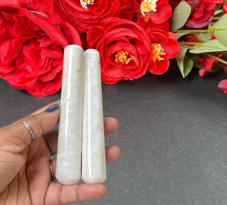 Puede incluir: Dos varitas de cristal cil&iacute;ndricas blancas con aspecto veteado. Las varitas se sostienen en una mano, con un fondo de vibrantes rosas rojas. Las varitas son lisas y pulidas, con una forma ligeramente c&oacute;nica.