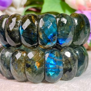 May include: A bracelet composed of faceted, rounded labradorite beads. The stones display a dark grey and olive green base color with iridescent flashes of electric blue and gold light reflecting from the polished surfaces of the natural mineral.