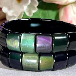 Könnte beinhalten: Ein Stapel aus zwei elastischen Armbändern, die aus glatten, quadratischen Perlen bestehen. Die Perlen sind überwiegend glänzend schwarz, wobei einzelne Perlen schimmernde, irisierende Akzente in Smaragdgrün, Limettengrün und tiefem Violett aufweisen.