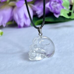 May include: Clear quartz crystal skull pendant on a black cord necklace. The skull pendant is intricately carved, showcasing the details of the skull's features. The pendant is suspended against a backdrop of purple flowers.