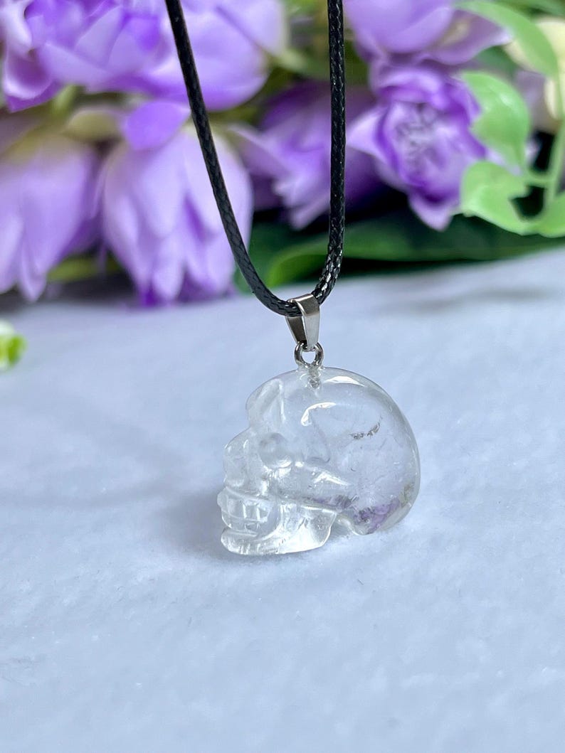 May include: Clear quartz crystal skull pendant on a black cord necklace. The skull pendant is intricately carved, showcasing the details of the skull's features. The pendant is suspended against a backdrop of purple flowers.
