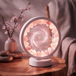 Peut inclure: Une lampe circulaire blanche dotée d'un anneau de cristaux de quartz rose brut et translucide entourant un affichage central lumineux représentant une galaxie en spirale. La lampe repose sur une base cylindrique blanche solide.