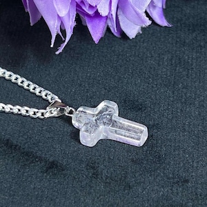 May include: A clear quartz crystal cross pendant on a silver chain. The cross is faceted and has a slightly translucent appearance. The chain is a curb style. The background is dark gray, with a purple flower in the upper left corner.