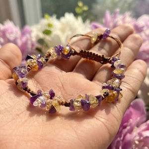 May include: A bracelet featuring small, irregular, polished chip beads in shades of purple amethyst and yellow citrine, strung on a dark brown cord. The design includes a sliding knot closure mechanism with two dangling ends finished with beads.