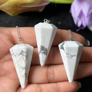 Puede incluir: Tres péndulos de piedras preciosas blancas y grises con cadenas plateadas. Los péndulos tienen forma de cono facetado. Las piedras preciosas tienen un aspecto veteado grisáceo. El fondo incluye una mano y flores moradas y amarillas.