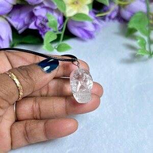 May include: A clear quartz crystal skull pendant on a black cord necklace. The skull is intricately carved with visible facial features. The background features purple and yellow flowers.