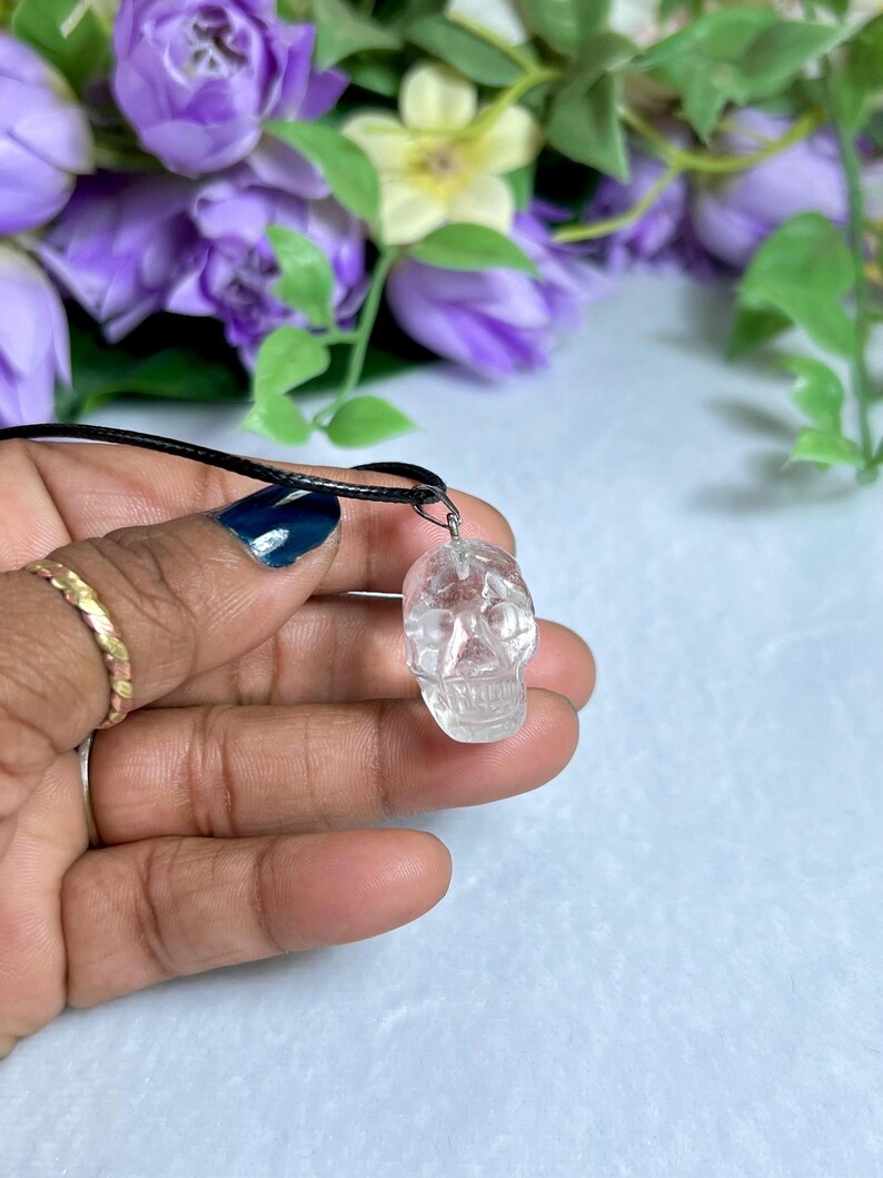 May include: A clear quartz crystal skull pendant on a black cord necklace. The skull is intricately carved with visible facial features. The background features purple and yellow flowers.