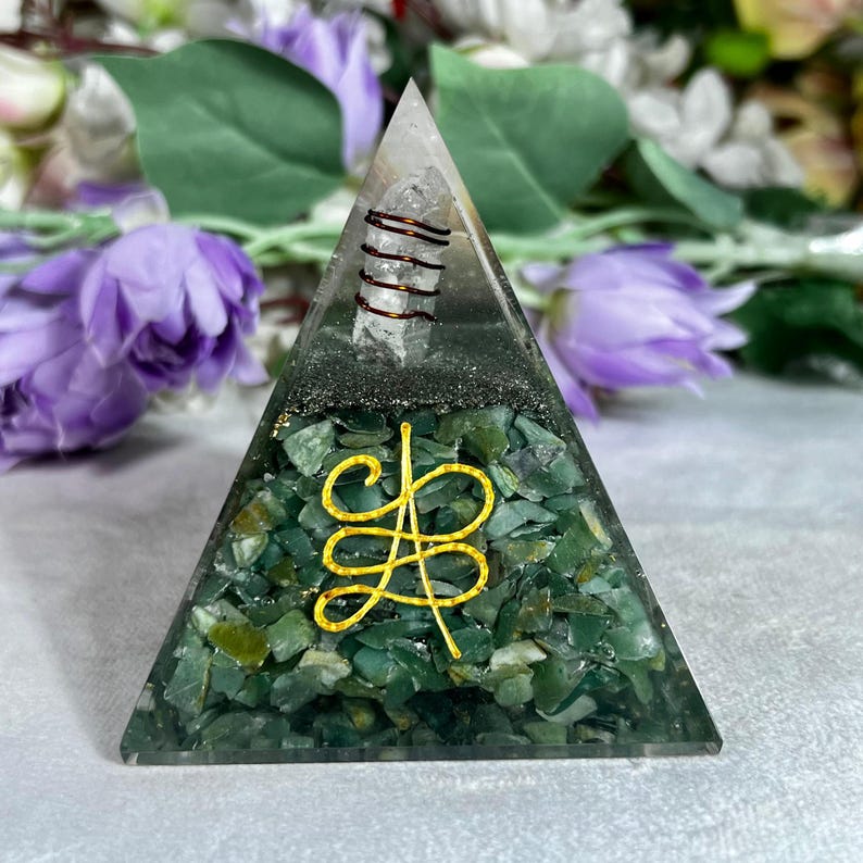 May include: A clear pyramid-shaped orgone crystal with a green base and a gold-colored symbol. The pyramid contains green stones, a clear crystal, and a copper coil. The background features purple flowers and green leaves.