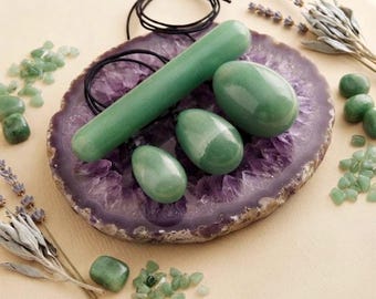 Puede incluir: Una varita de aventurina verde y tres huevos de aventurina verde, cada uno con un cordón negro, dispuestos sobre una gran rodaja de geoda de amatista morada. Pequeñas virutas de aventurina verde y ramas de lavanda seca están dispersas.