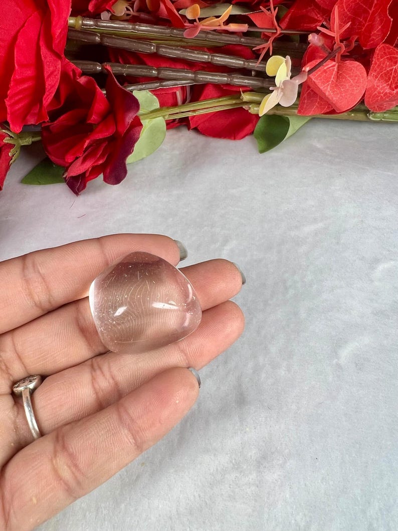 May include: A clear, rounded crystal stone held in a hand. The stone has a smooth, polished surface and a slightly pinkish hue. Red flowers and greenery are visible in the background, adding a touch of color.