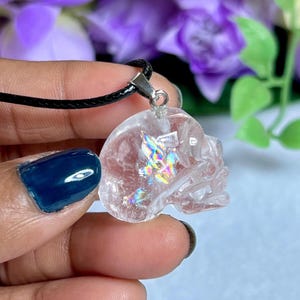 May include: Clear quartz crystal skull pendant on a black cord. The skull pendant has iridescent inclusions that catch the light. The pendant is attached to a silver-colored bail.