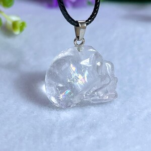 May include: A clear quartz crystal skull pendant on a black cord necklace. The skull is intricately carved with visible details and iridescent inclusions. The pendant hangs from a silver-colored bail.