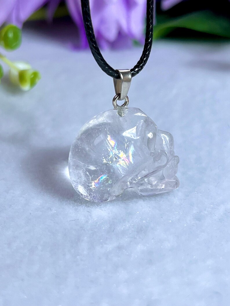 May include: A clear quartz crystal skull pendant on a black cord necklace. The skull is intricately carved with visible details and iridescent inclusions. The pendant hangs from a silver-colored bail.