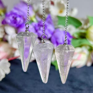 Puede incluir: Tres péndulos de cristal transparente facetado que cuelgan de cadenas de metal plateado. Cada péndulo presenta una forma geométrica puntiaguda y multifacetada con un brillo iridiscente reflectante que capta la luz, creando sutiles reflejos púrpuras y blancos.